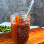 A head on image of a mason jar filled with homemade bbq sauce on a wooden board with a spoon in the jar and drips of sauce on the side