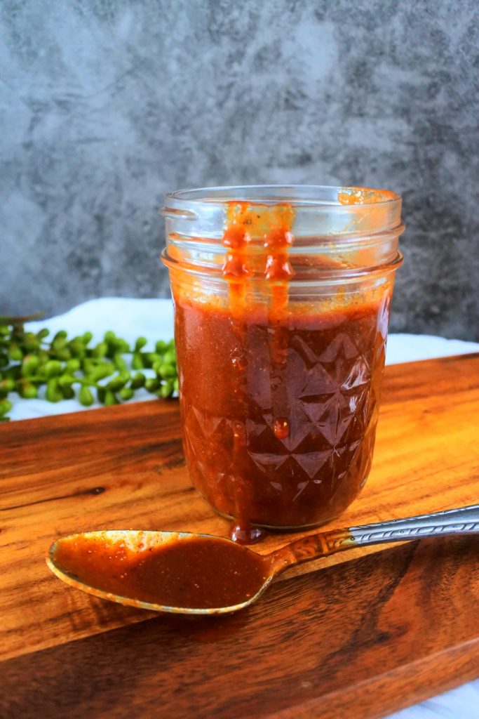 A head on image of a mason jar filled with homemade bbq sauce on a wooden board with a spoon with sauce on the board and drips of sauce running down the jar