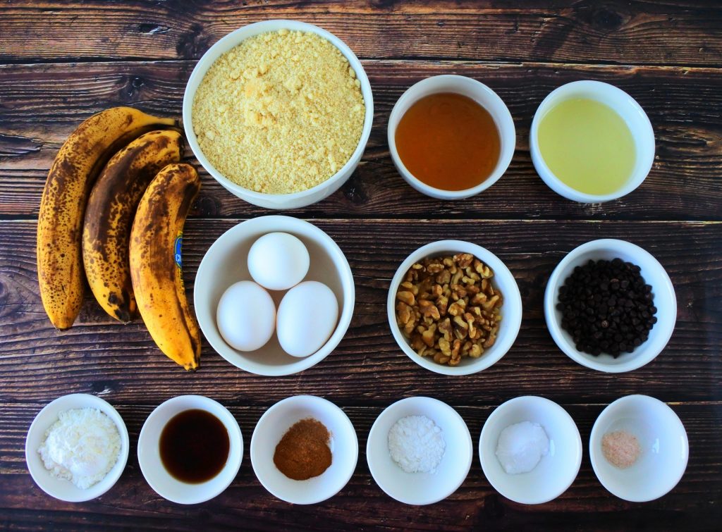 An overhead image of ingredients for a banana nut bundt bread including overripe bananas, blanched almond flour, honey, oil, eggs, walnuts, mini dark chocolate chips, salt, baking soda, baking powder, cinnamon, vanilla extract and potato starch