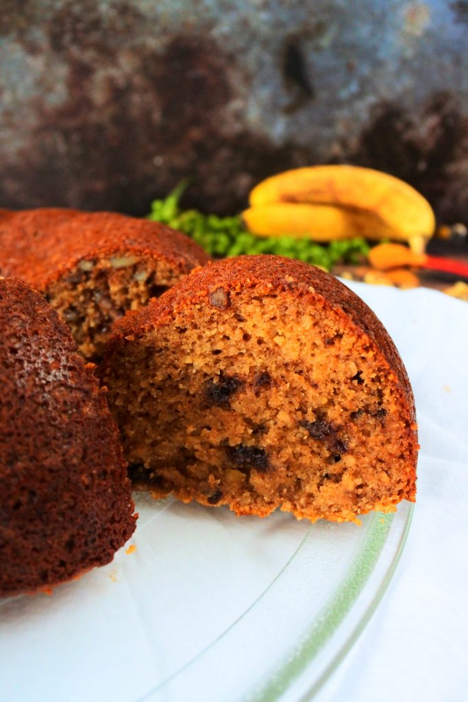 A close up image of a banana nut bundt bread with a piece sliced out of it with banana, nuts, chocolate and spices in the background