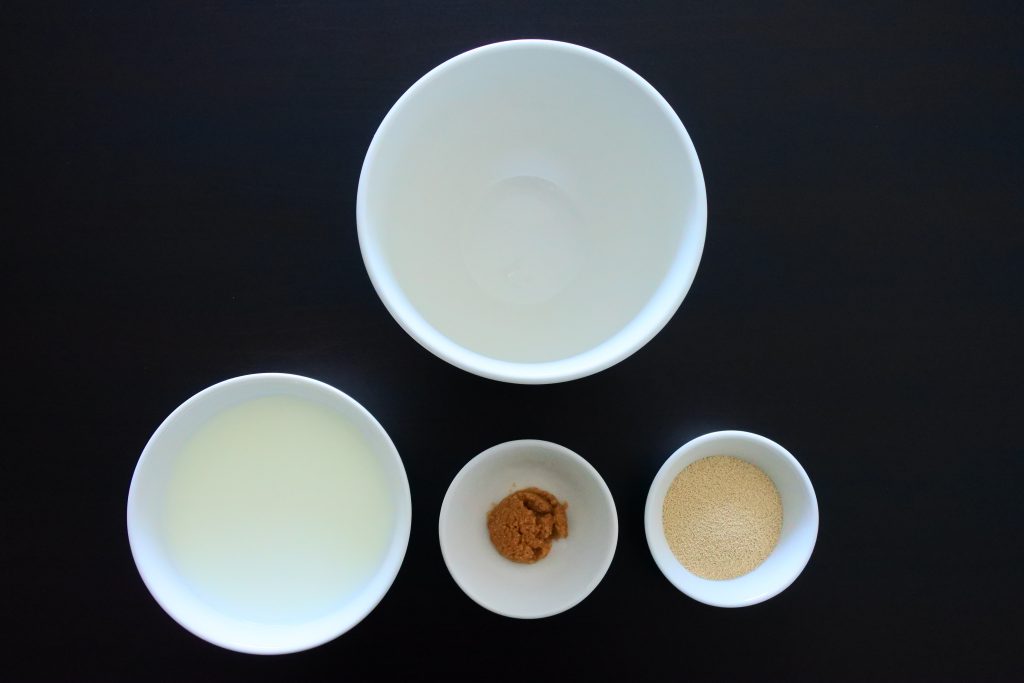 An overhead image of four bowls, one larger empty one, one containing milk, another containing sugar and the last containing yeast