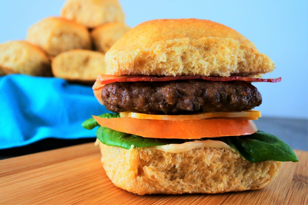 A close up image of a slider with bacon, a beef patty, cheese, tomato, spinach and mayo on a Hawaiian sweet dinner roll with a basket of rolls in the background