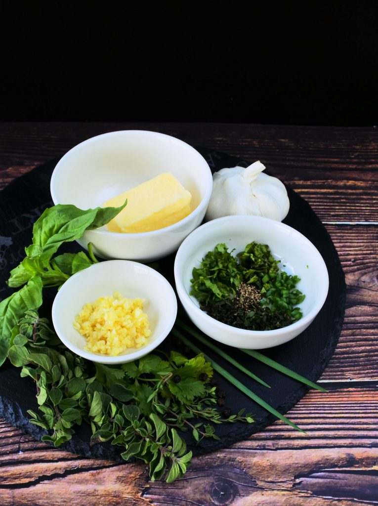 An angled image of a slate plate containing dishes of diced garlic, assorted chopped fresh herbs, softened butter, all surrounded by fresh herbs and spices
