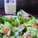 A head on image of a dressed Caesar salad with a bottle of homemade Caesar Salad Dressing in the background.