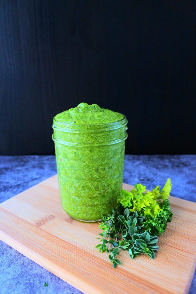A head on image of green seasoning in a jar with a bunch of fresh herbs at its base