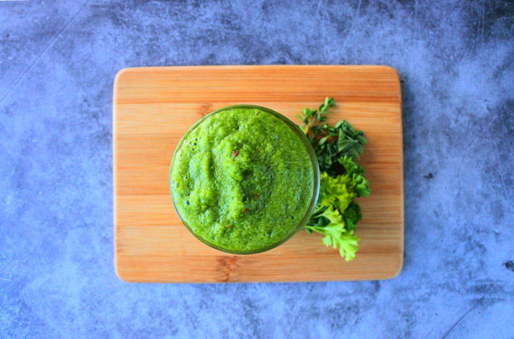An over head image of a jar of fresh green seasoning on a wooden board with fresh herbs at the base