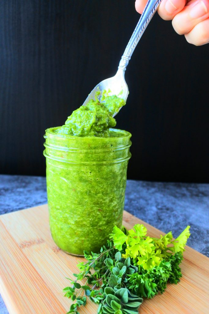 A head on image of green seasoning in a jar with a spoonful being lifted out and a bunch of fresh herbs at its base
