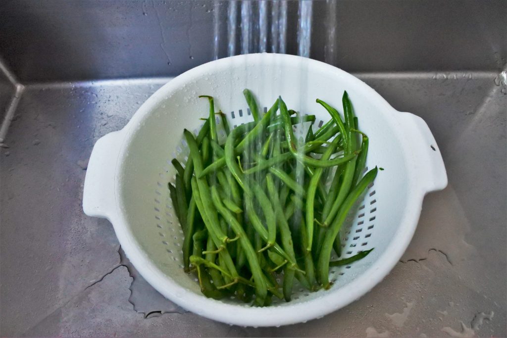 An angled image of a colander filled with green beans in a sink with a stream of water over it.