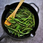An overhead image of a cast iron skillet of charred green beans with a wooden utensil in it