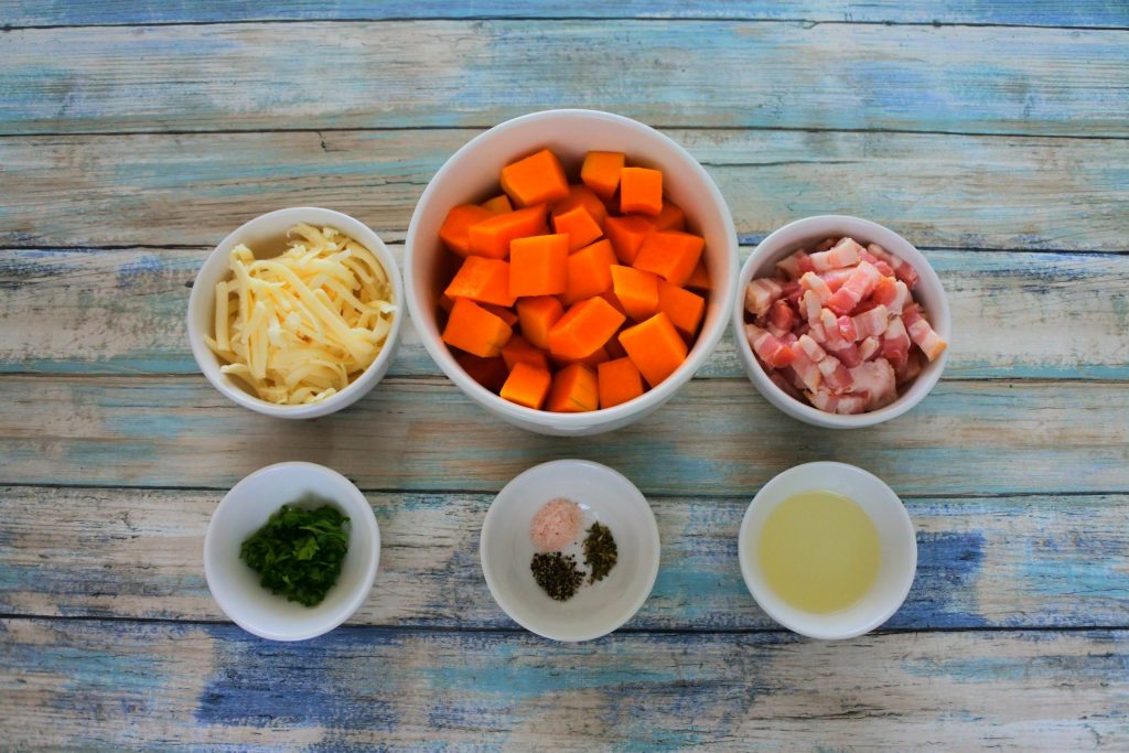 An overhead image of bowls of ingredients including shredded cheese, cubed butternut squash, bacon pieces, light olive oil, salt, black pepper and oregano, and fresh parsley