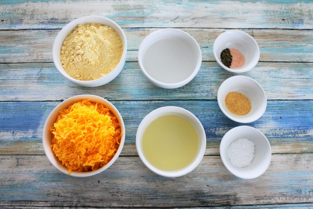 An overhead image of bowls of ingredients for cheesy chickpea crackers including: chickpea flour, shredded cheddar cheese, water, light olive oil, salt, pepper, garlic powder and baking powder