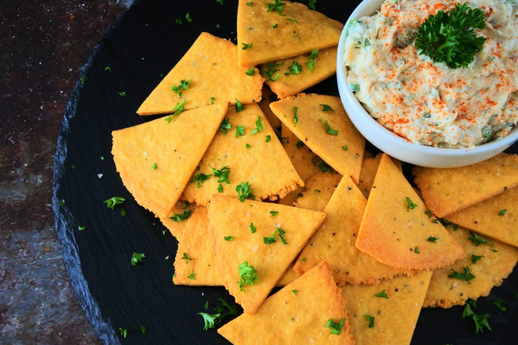 An overhead image of a plate of cheesy chickpea crackers topped with coarse pink salt and fresh parsley with a small dish of creamy dip