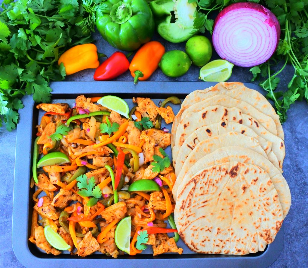 An overhead image of cooked chicken fajitas next to a row of soft and fluffy whole wheat tortillas on a sheet tray with an assortment of fresh veggies to the top border