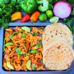 An overhead image of cooked chicken fajitas next to a row of soft and fluffy whole wheat tortillas on a sheet tray with an assortment of fresh veggies to the top border
