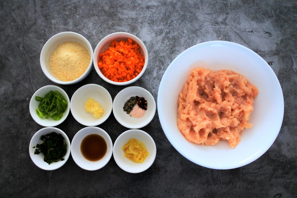 An overhead view of bowls of ingredients for Thai Chicken Meatballs including ground chicken, diced carrots, almond flour, sliced chives, grated garlic, salt, pepper, red pepper flakes, fresh cilantro, fish sauce, and grated ginger
