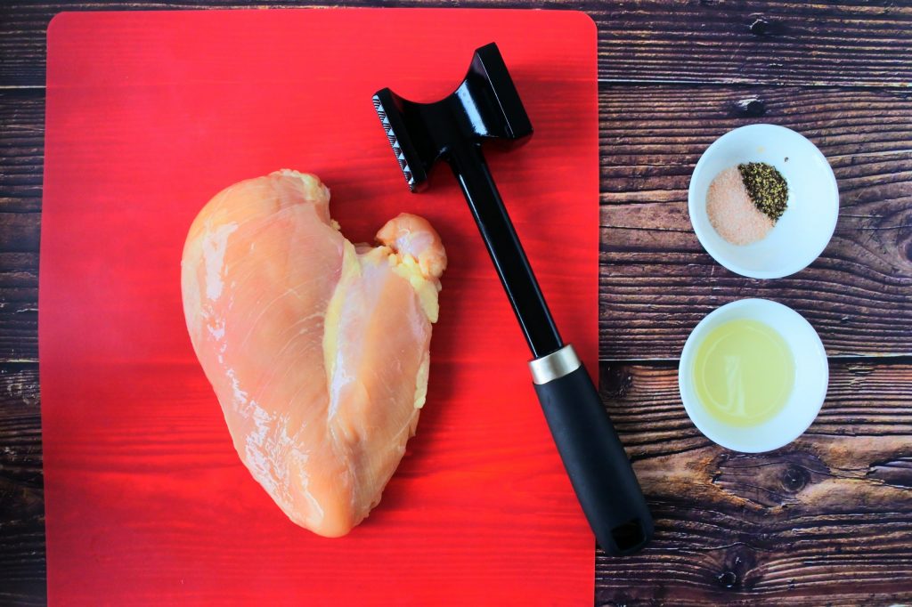 An overhead image of a raw chicken breast on a cutting mat next to a meat mallet with two small dishes of oil and salt and pepper on the side.