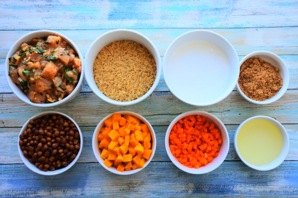An overhead image of bowls containing ingredients for pelau including seasoned cubed chicken, rice, coconut milk, raw brown sugar, pigeon peas, cubed squash, diced carrots and light olive oil