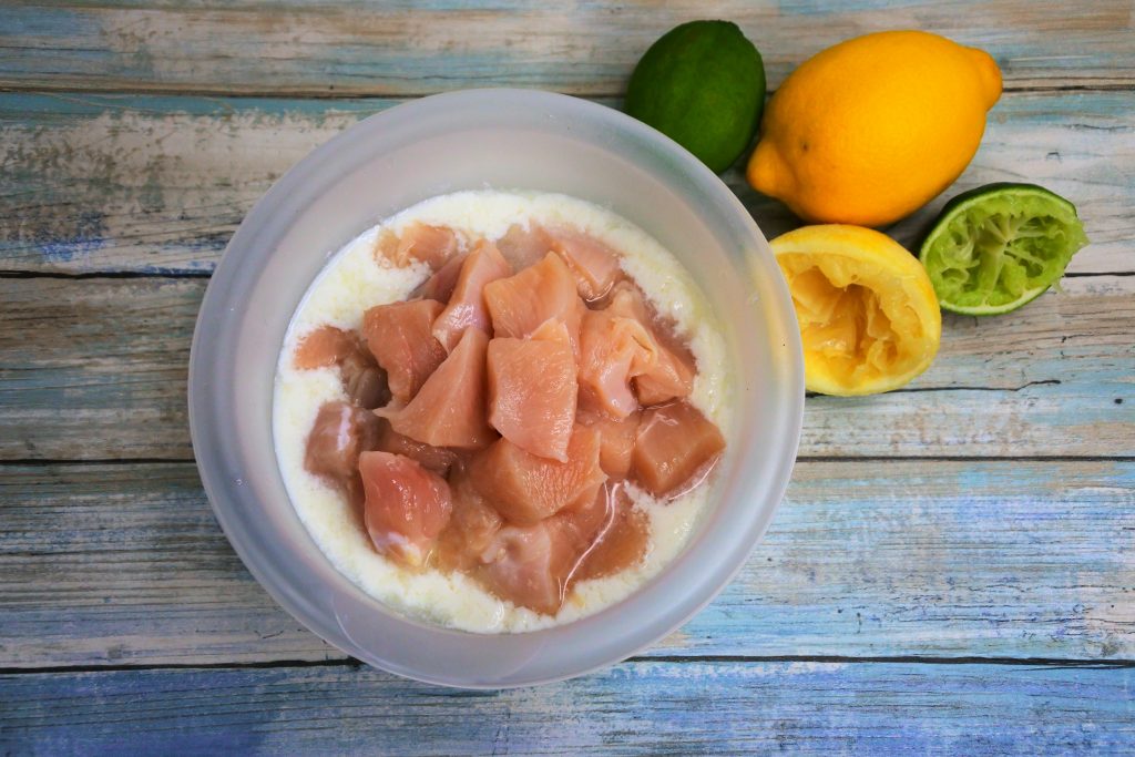 An overhead image of a bowl of cubed chicken pieces in a buttermilk marinade with fresh limes and lemons in the top right corner