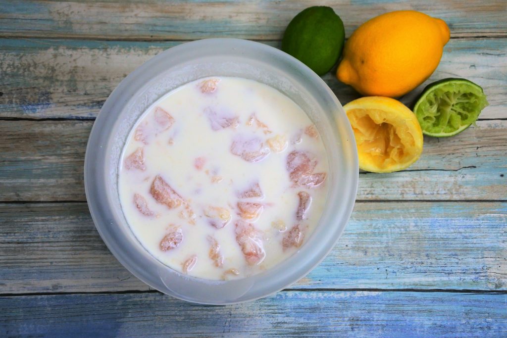 An overhead image of a bowl of cubed chicken pieces mixed in a buttermilk marinade with fresh limes and lemons in the top right corner