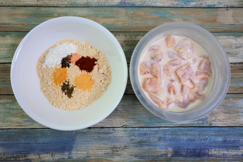 An overhead image of a bowl of whole wheat flour and spices next to a bowl of cubed chicken pieces in a buttermilk marinade