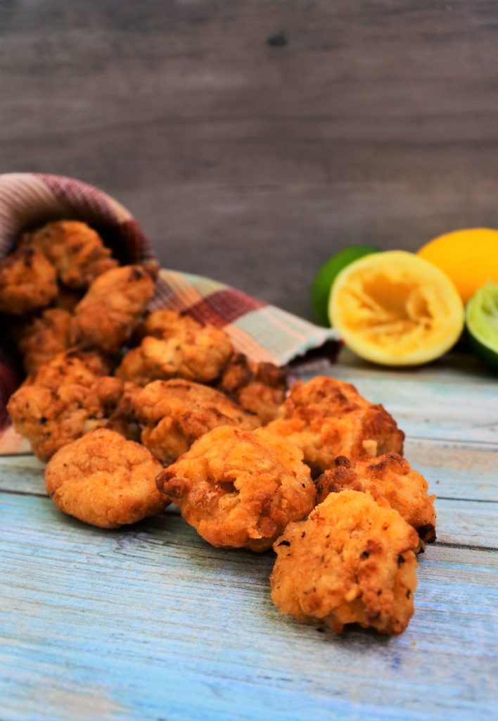 A vertical head on image of some popcorn chicken spilling out of a linen cone with a some fresh limes and lemons in the background