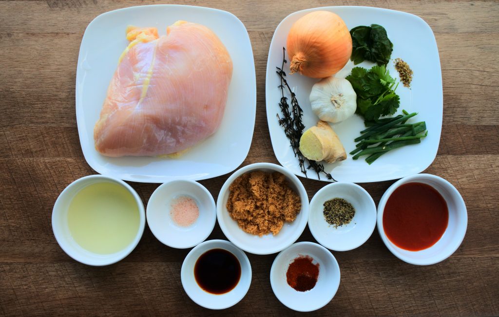 An overhead image of various plates of dishes of ingredients including: chicken, salt, pepper, gluten-free soy sauce, paprika, chipotle powder and cayenne pepper, tomato sauce, thyme, onion, garlic, chives, ginger, basil, cilantro, dried oregano, oil, and sugar.