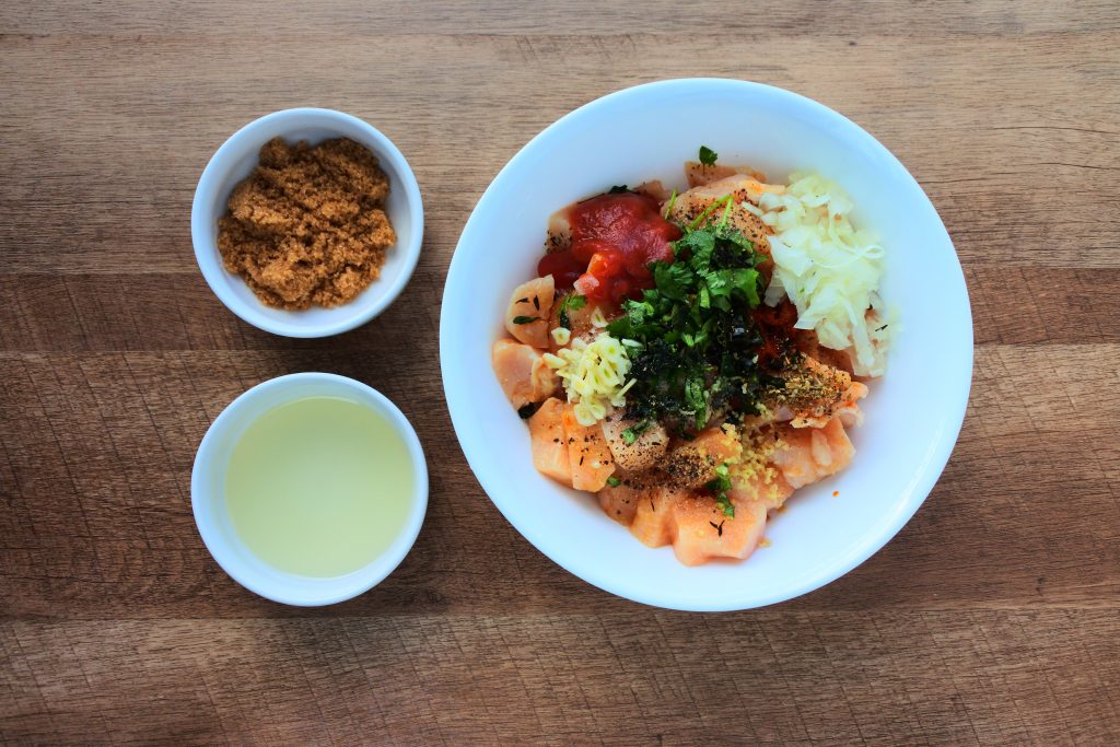 An overhead image of raw cubed chicken topped with various herbs and spices with two dishes on the left containing raw organic brown sugar and light olive oil