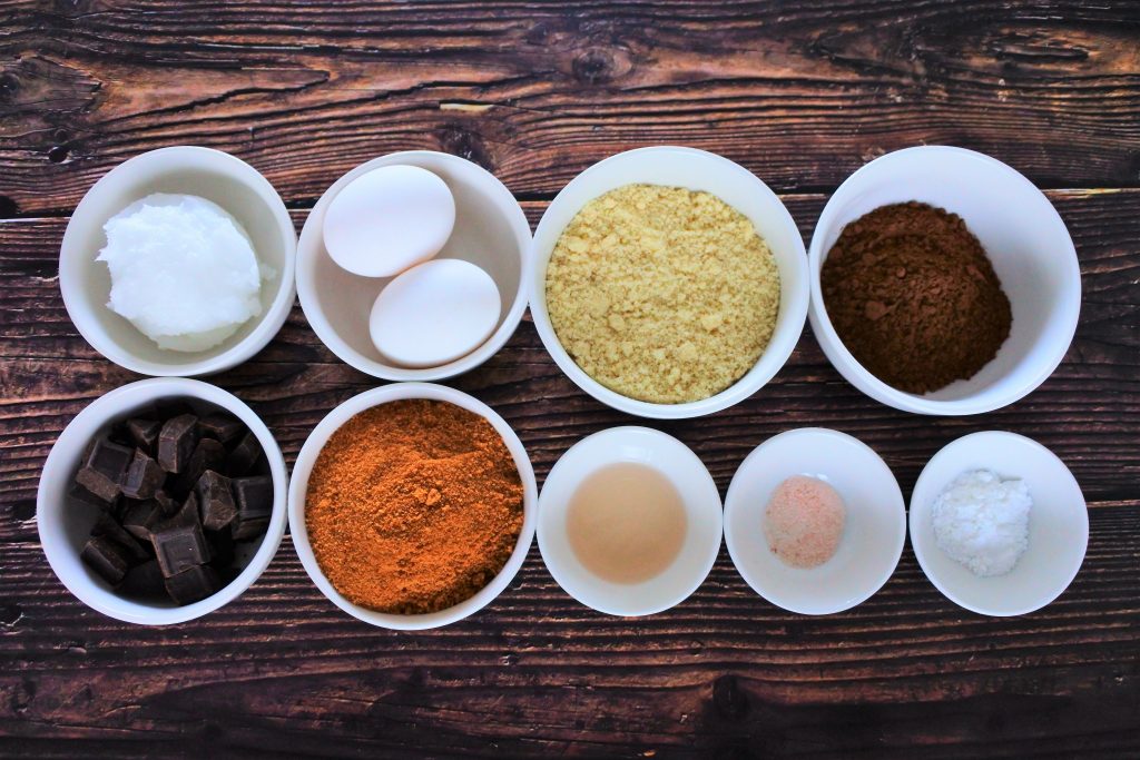 An overhead image of bowls containing ingredients for clean eating brownies including Dark chocolate, coconut oil, eggs, coconut sugar, almond flour, cocoa powder, baking powder, salt, and vanilla extract.