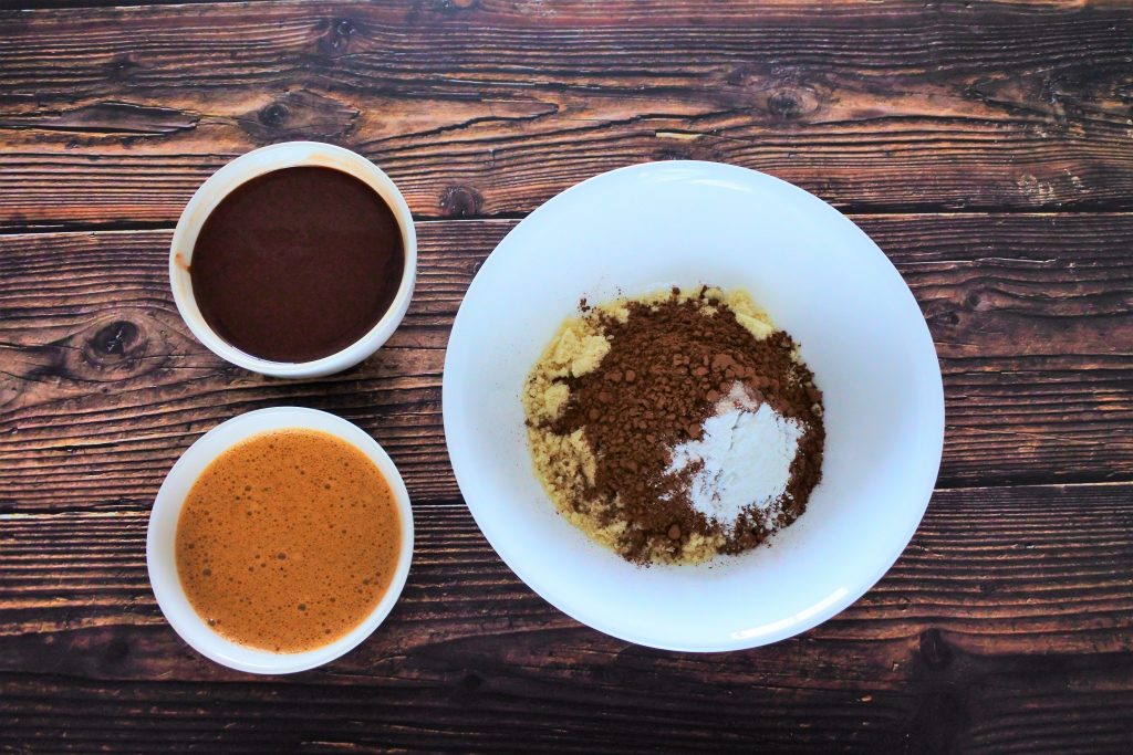 An overhead image of three bowls, one containing uncombined dry ingredients including almond flour, cocoa powder, salt and baking powder, another bowl containing a ,ix of melted chocolate and coconut oil, and the third containing a blend of eggs and coconut sugar