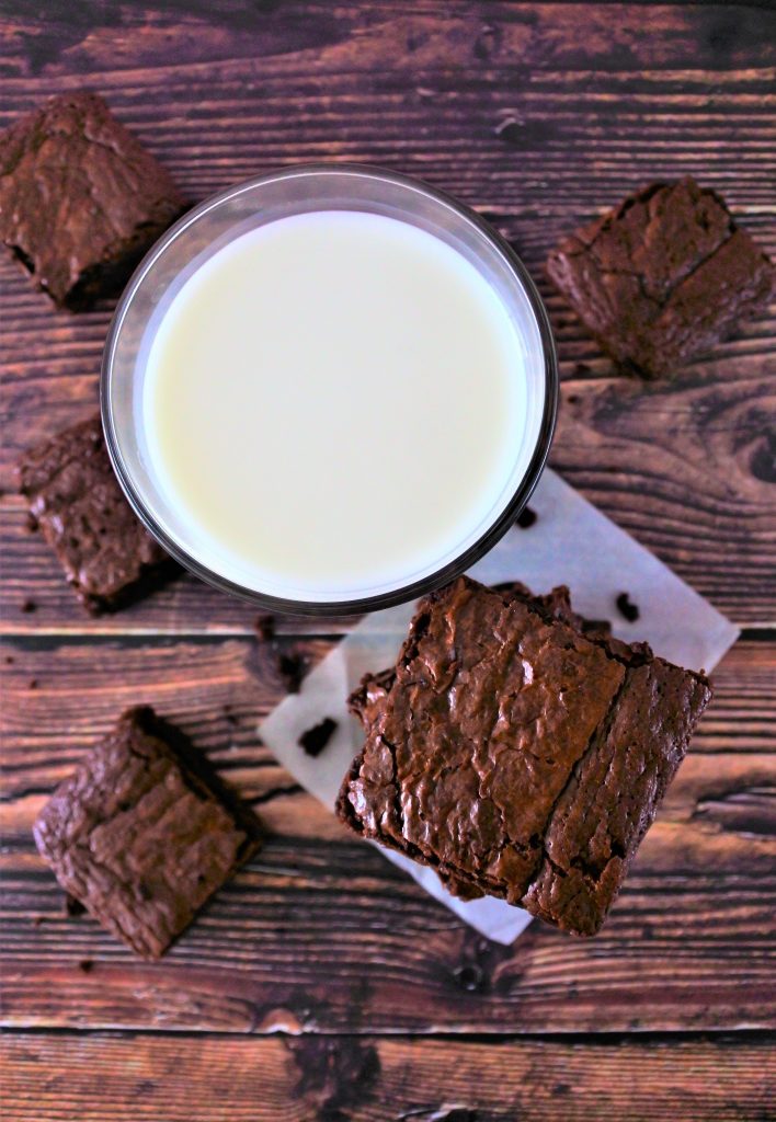 Am overhead image of a glass of milk and a tower of brownies surrounded by squares of brownies on a wooden tabletop