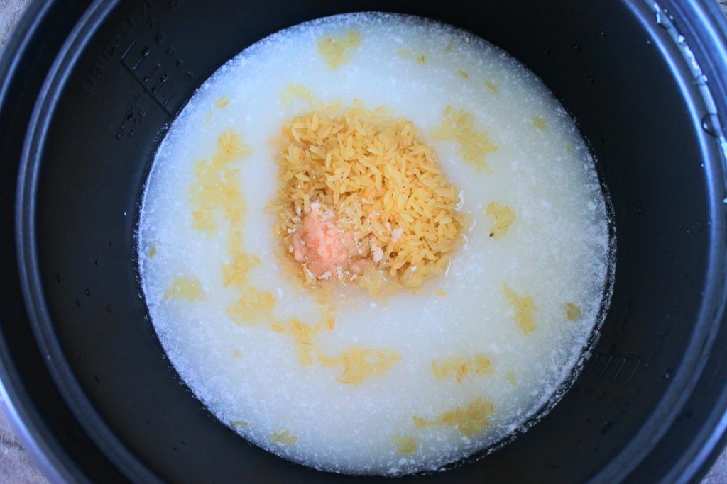 An overhead image of a rice cooker with coconut milk, dry rice and salt