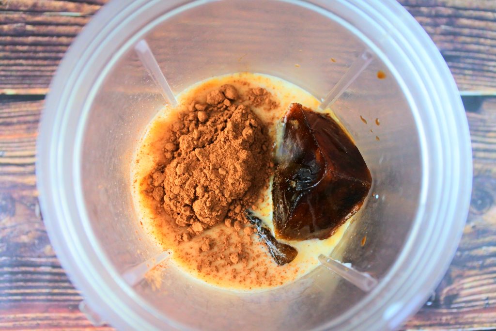 An overhead image of ingredients for a blended iced coffee frappe in a cup including milk, coffee ice cubes and cocoa powder