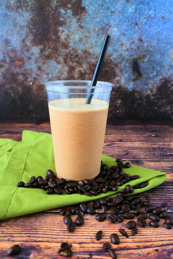 A head on image of a blended iced coffee frappe with a black straw on a green linen napkin surrounded by coffee beans.