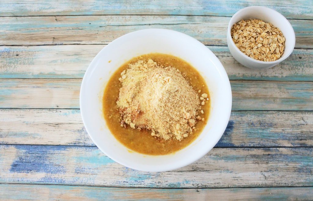 An overhead image of a bowl of a mashed banana mixture with almond flour added in and a smaller bowl containing rolled oats on the side