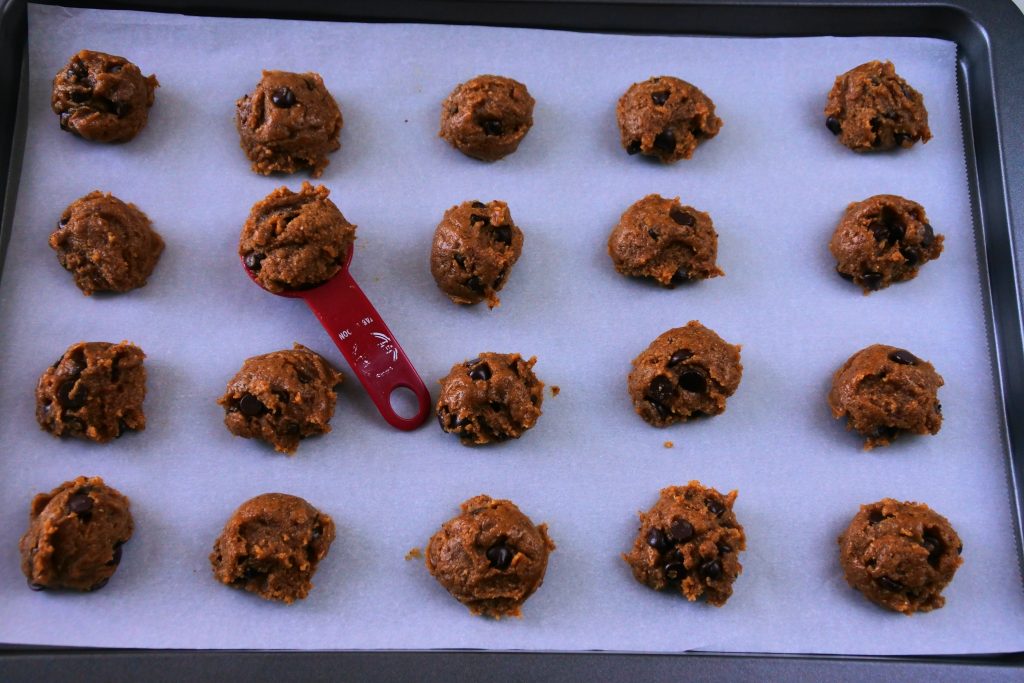 A tilted overhead image of a tray of twenty unbaked chocolate chip cookies with a red measuring spoon containing a portion of chocolate chip cookie dough in the second row of the tray