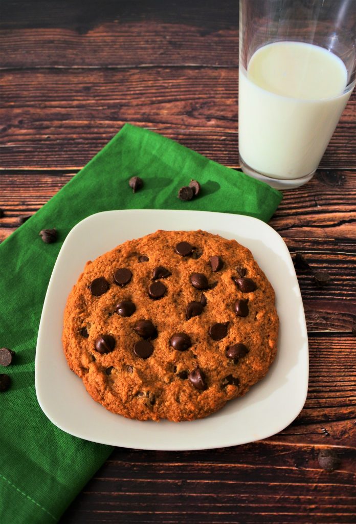A tilted angle overhead image of a large chocolate chip cookie on a plate surrounded by a scattering of chocolate chips, a green napkin and a glass of milk.