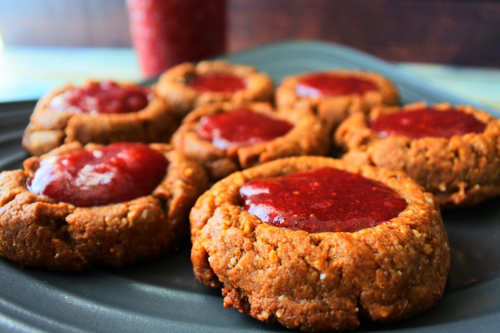 A close up image of a PB&J thumbprint cookies on a plate with a jar of homemade strawberry jam in the background