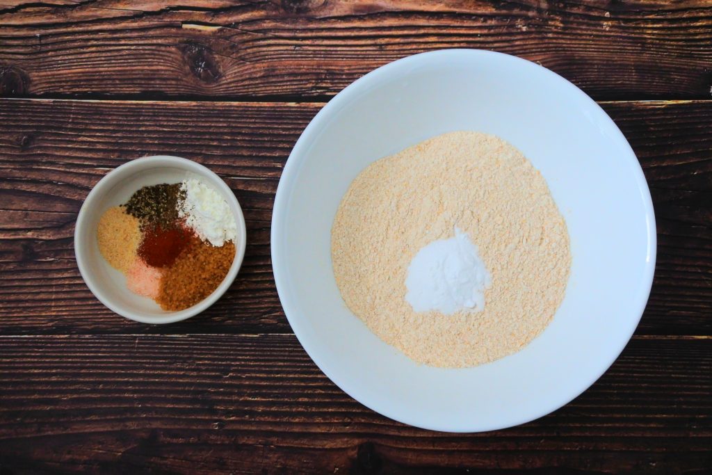 An overhead image of a bowl of flour with some baking powder next to a dish containing a spice mix