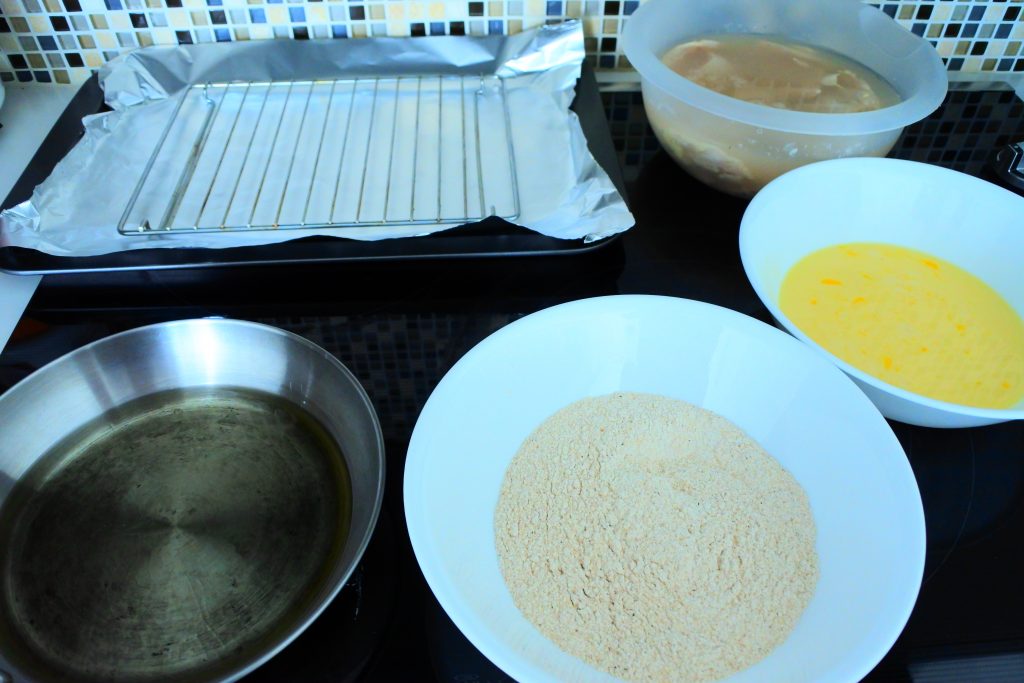 An angled image of the set up for frying chicken including a frying pan with oil, a bowl of flour mixture, a bowl of egg wash, a bowl of chicken pieces and a wire rack on a sheet of foil paper