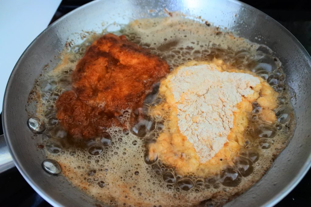 A close up image of flour and egg wash dredged chicken pieces frying in a frying pan