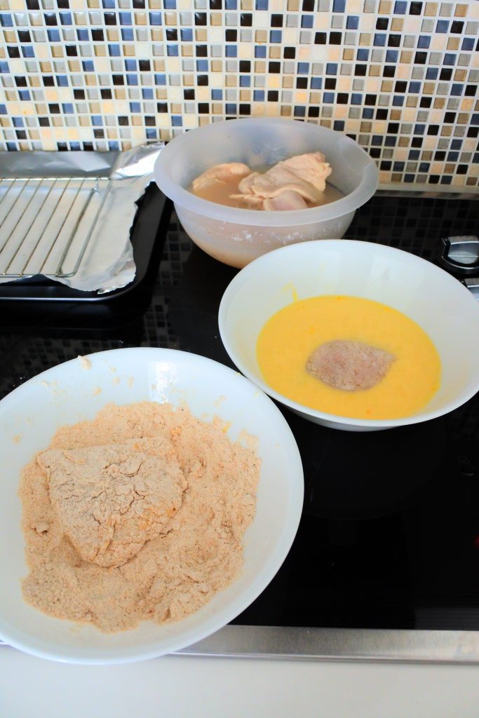 An angled image of a prep station for frying chicken including a bowl with a flour mix, a bowl with an egg wash, a bowl with chicken pieces and a wire rack on foil paper