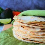 A close up head on image of a stack of freshly made corn tortillas wrapped in a green linen napkin with fresh produce in the background