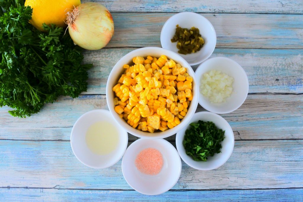 An overhead of bowls of ingredients for corn salsa including fresh corn, jalapeno, onion, cilantro and parsley, salt and lemon juice