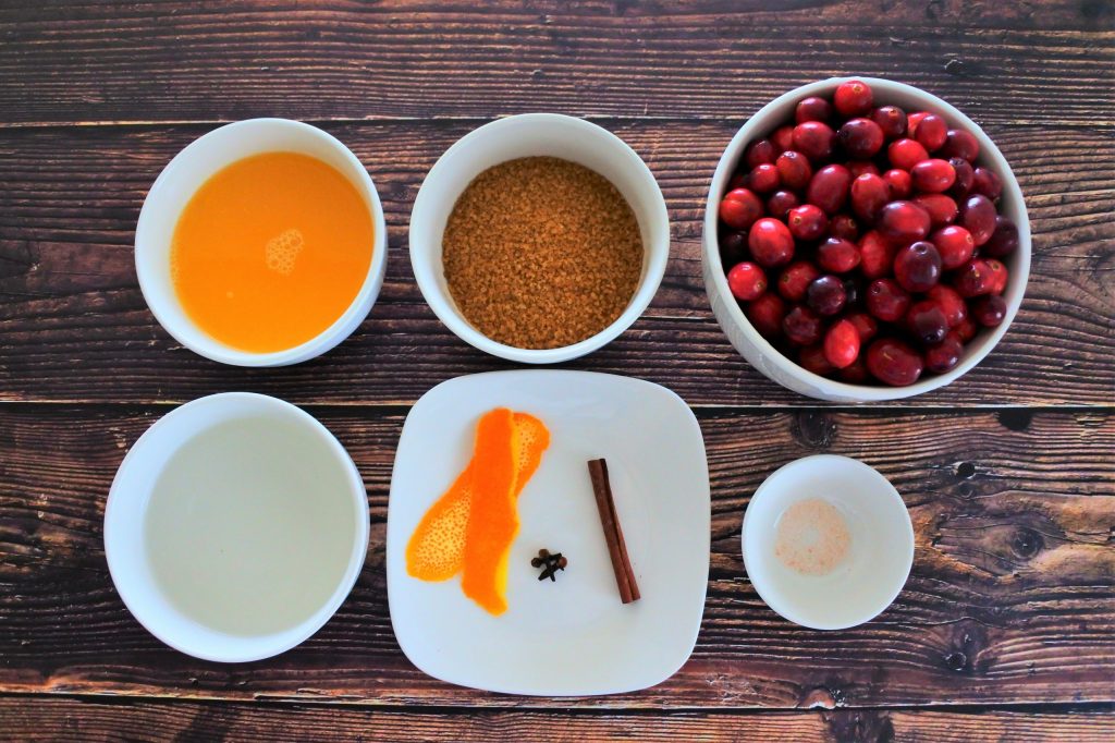 An overhead image of ingredients for homemade cranberry sauce including freshly squeezed orange juice, raw cane sugar, fresh cranberries, water, orange peel, whole cloves, a stick of cinnamon and salt.