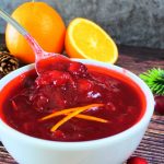 An angled image of a bowl with a spoon lifting out some homemade cranberry sauce garnished with orange peel slivers surrounded by fresh oranges, pine cones, a cinnamon stick and fresh cranberries