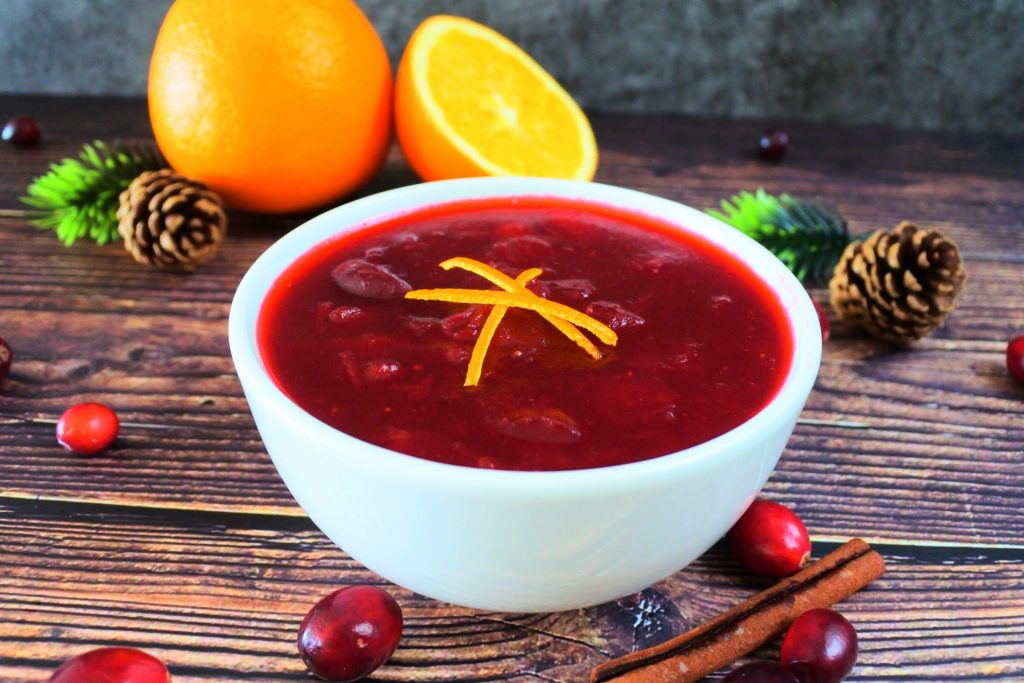 An angled image of a bowl of homemade cranberry sauce garnished with orange peel slivers surrounded by fresh oranges, pine cones, a cinnamon stick and fresh cranberries