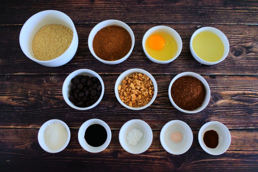 An overhead image of bowls containing ingredients for Double Chocolate Hazelnut cookies including almond flour, coconut sugar, an egg, oil, dark chocolate chips, chopped toasted hazelnuts, cocoa powder, coconut flour, molasses, baking powder, salt and espresso powder