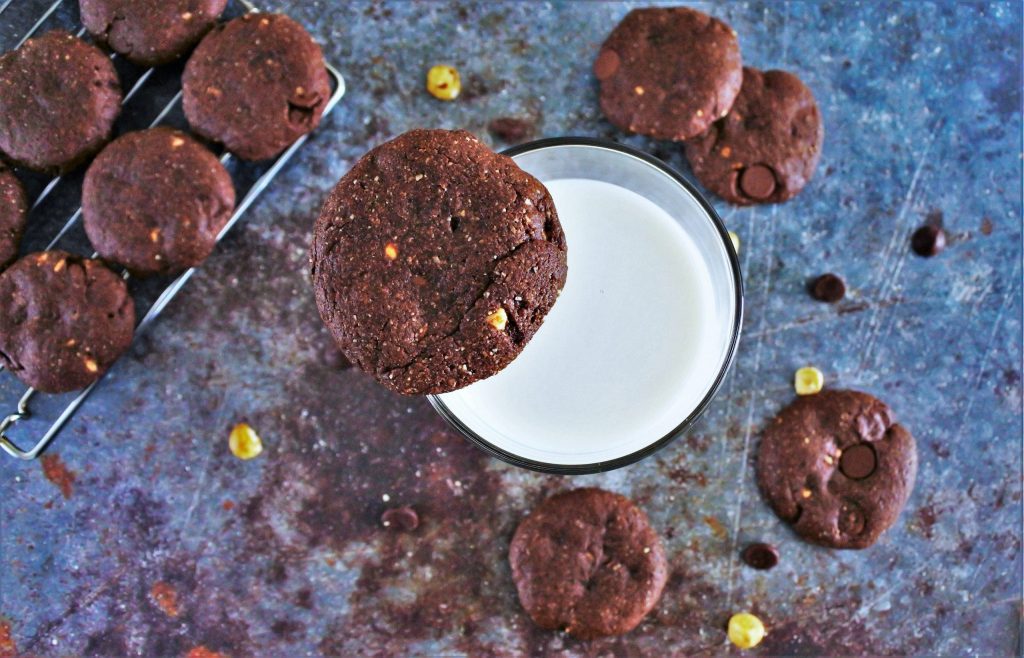 An overhead image of a glass of milk with a Double Chocolate Hazelnut Cookie balanced on the rim of the glass surrounded by other cookies