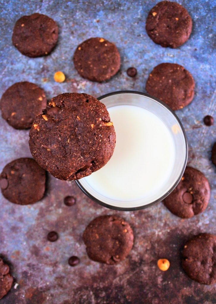 An overhead image of a glass of milk with a Double Chocolate Hazelnut Cookie balanced on the rim of the glass surrounded by other cookies