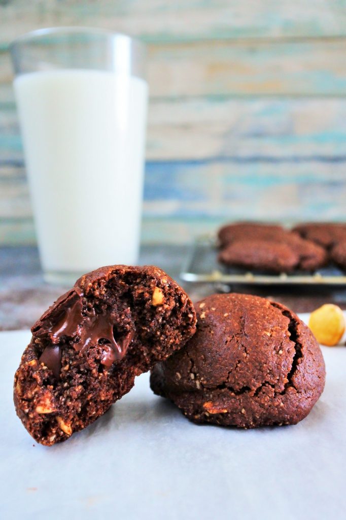 A head on image of a whole and a half domed Double Chocolate Hazelnut Cookie on parchment with a glass of milk and a wire rack with cookies on it in the background